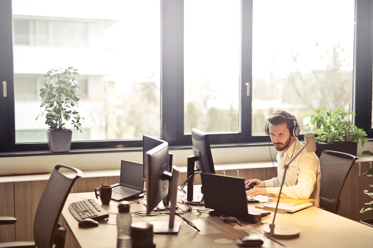 Services-01 A businessman sits at a desk using multiple computers and a headset in a well-lit modern office.